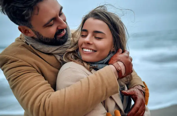 Hombre abrazando con ternura a una mujer mientras ambos sonríen en una playa ventosa